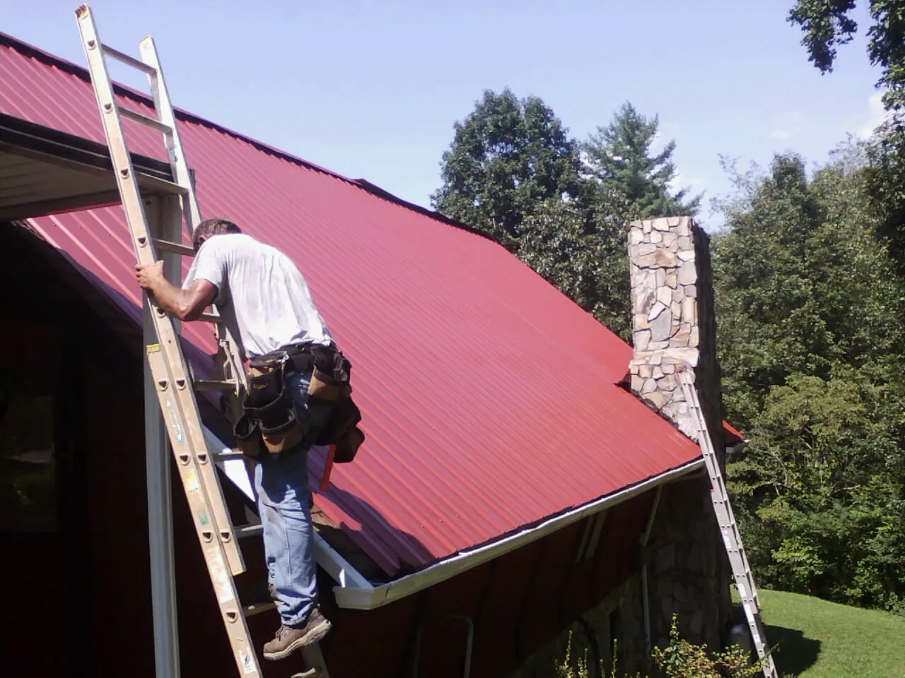 Roofer on a ladder beside a red metal roof and stone chimney — Soffit & Fascia Repair by Roofing Brothers in Dacula